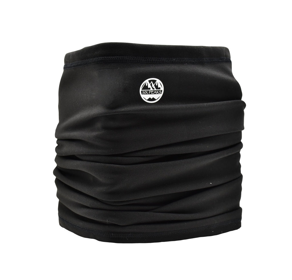 The Six Peaks Winter Neck Warmer is black with a small white mountain logo and "SIX PEAKS" text on the front. Its soft, slightly gathered fabric is wind resistant for added comfort in cold weather.