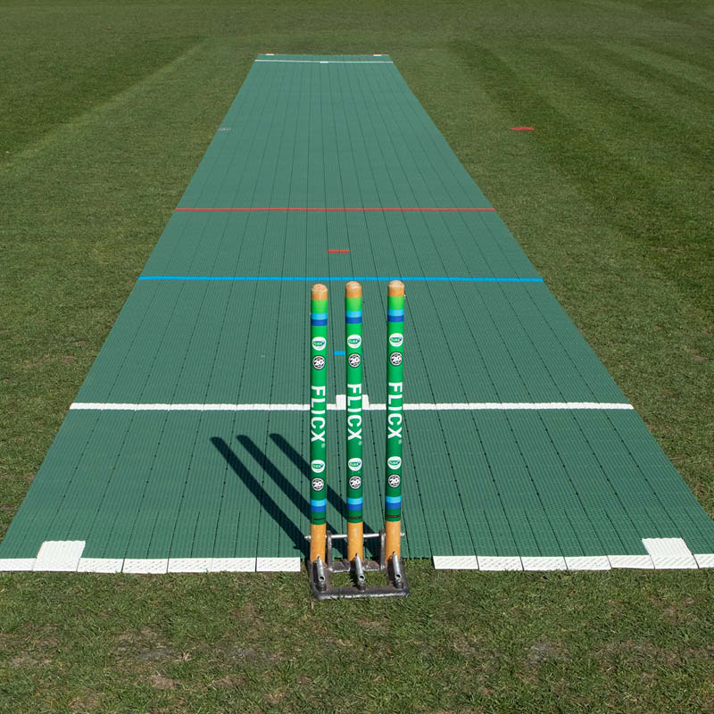 Three cricket stumps stand on the Flicx UK Flicx 2G MAP (Multi Age Pitch) Match Cricket Pitch, featuring white and colored lines on a green mat—perfect for clubs and schools needing versatile pitch setups.