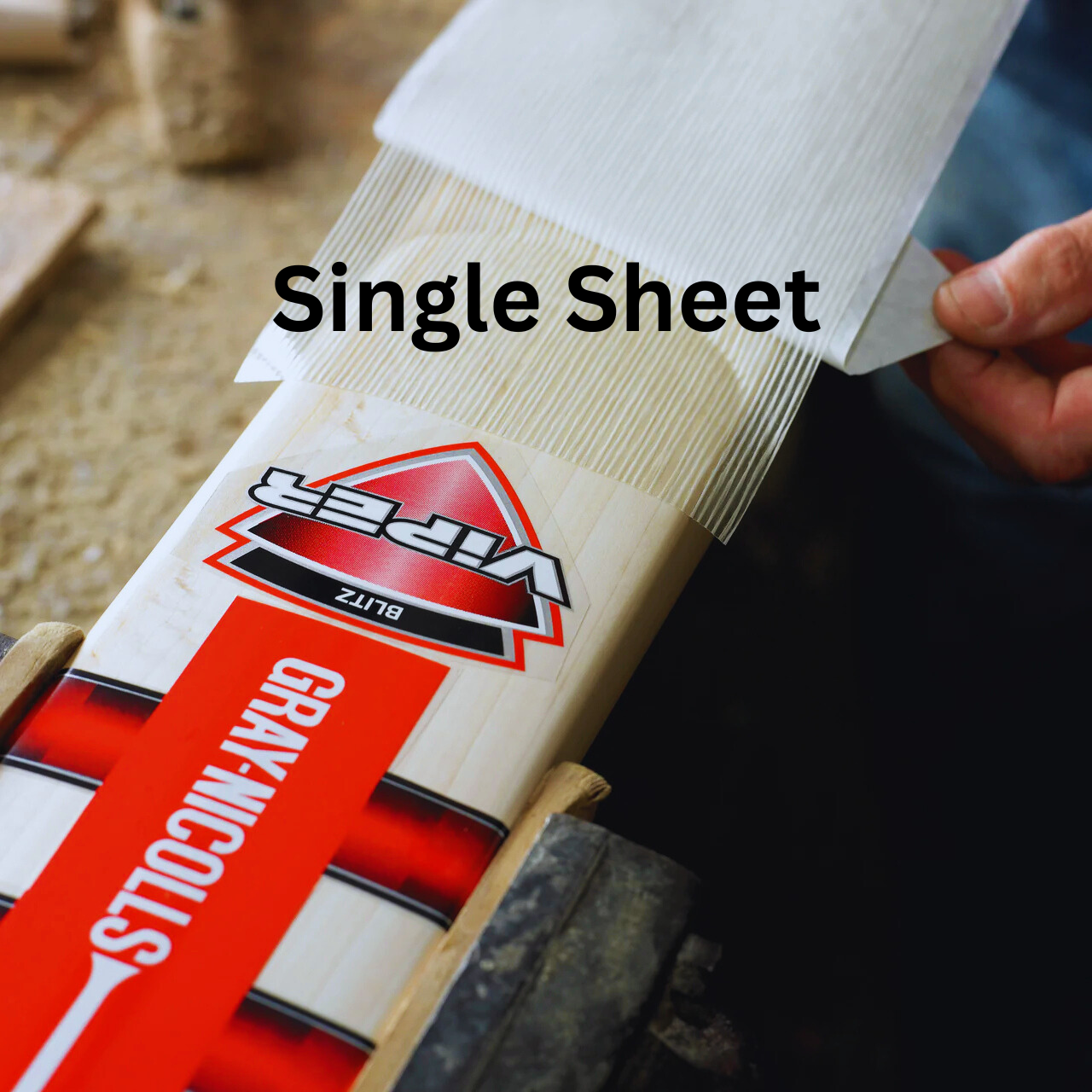 A hand applies a transparent Fibretec Sheet by Gray Nicolls to a Viper Blitz cricket bat on a workbench, providing superior bat protection.