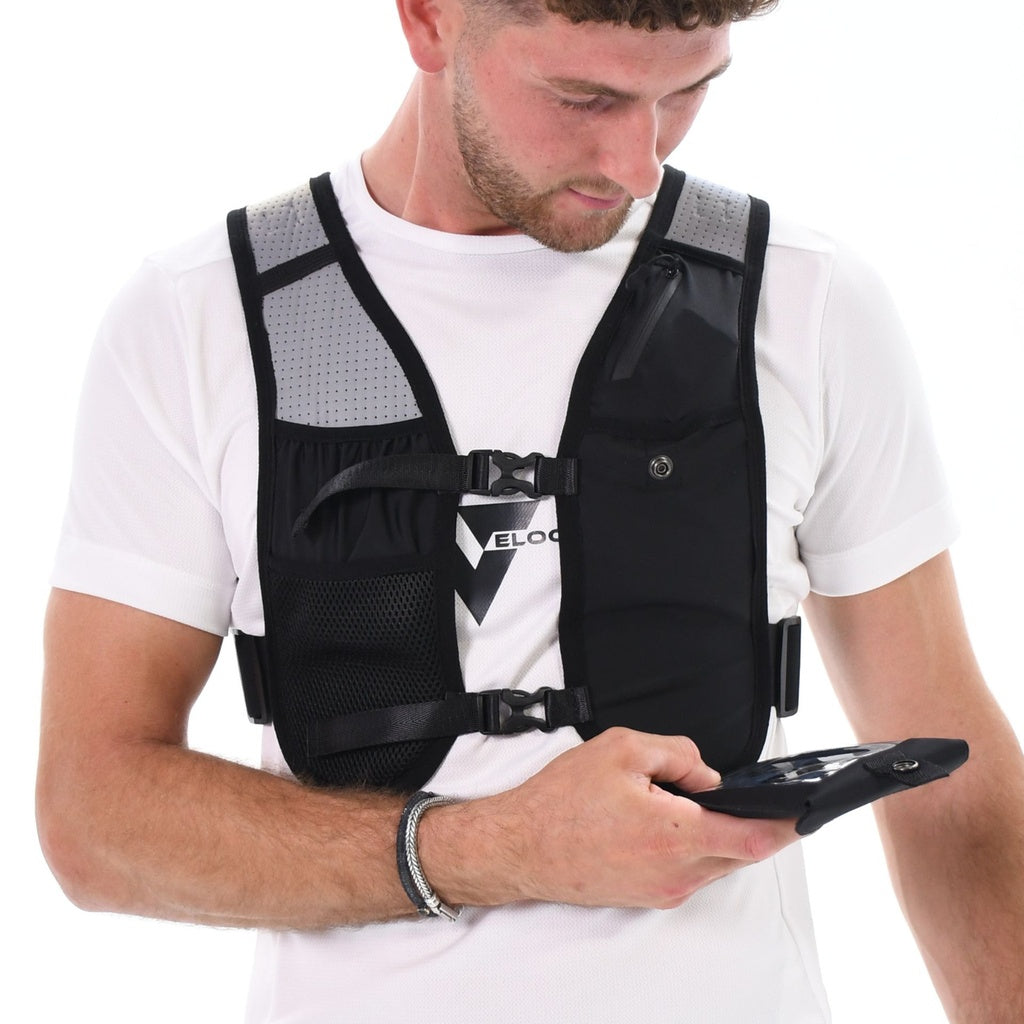 A man wearing a white t-shirt and the black Six Peaks Hydration Running Vest with mesh pockets looks down at a smartphone in his left hand. The background is white.