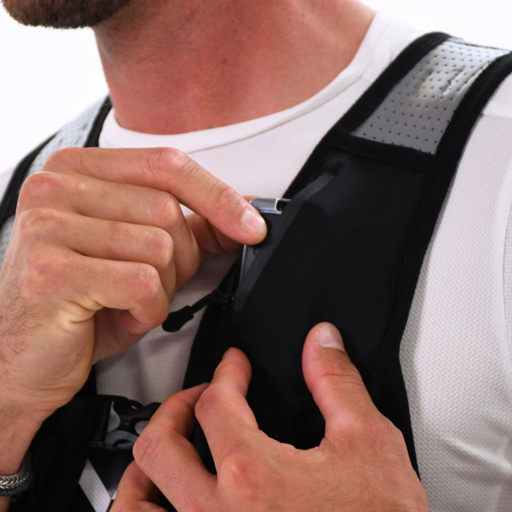 A close-up shows a person adjusting the zipper on the front of a Six Peaks Hydration Running Vest in black with gray details, worn over a white shirt. Only their hands, torso, and lower face are visible.