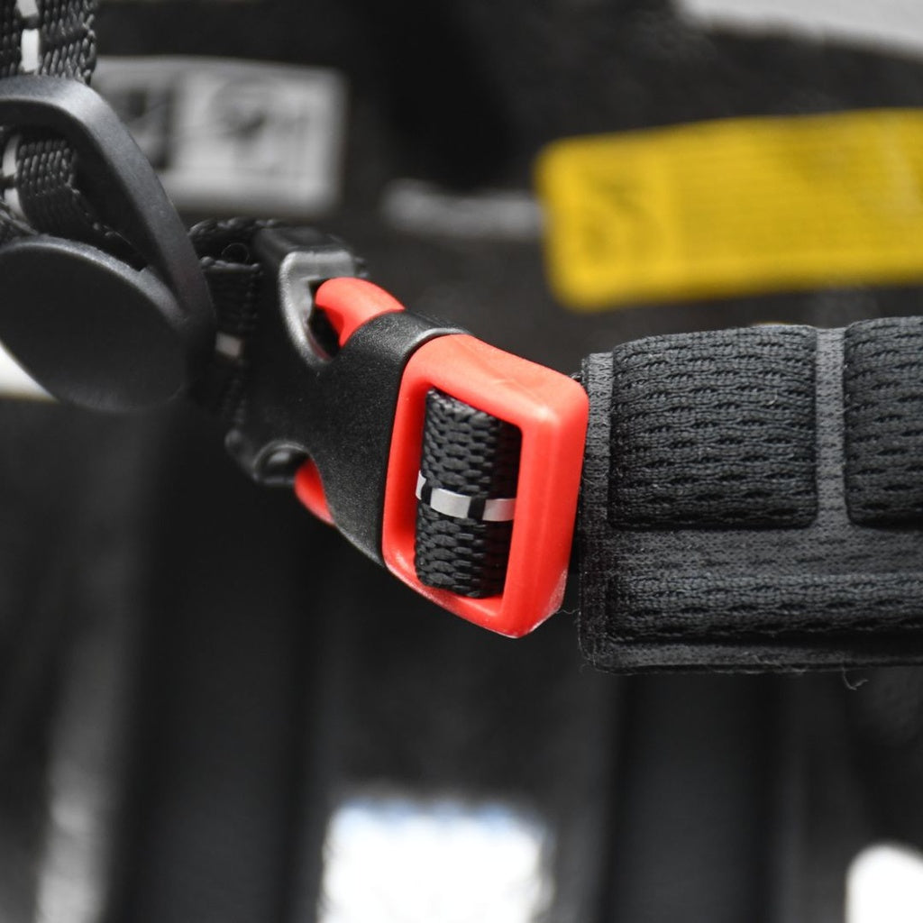 Close-up of a black and red plastic buckle fastened to a black strap with visible stitching on the Six Peaks Junior Cycling Helmet by Six Peaks. A blurred yellow label appears in the background.