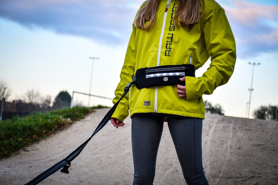 A person stands outdoors on a dirt path wearing the Six Peaks LED Dog Lead Running Belt by Six Peaks, which features a hands-free waist pouch. The sky is partly cloudy with trees and light poles visible in the background.