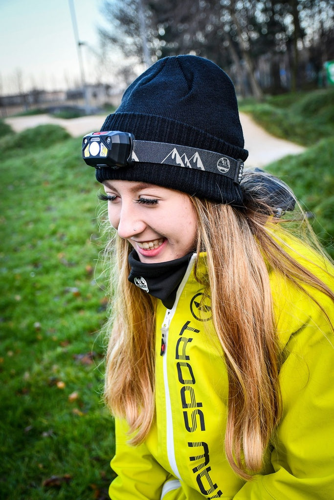 A young woman with long blonde hair, wearing a black beanie featuring the Six Peaks LED Head torch with Motion Sensor, a yellow jacket, and a black neck gaiter, smiles while sitting outdoors on grassy ground.