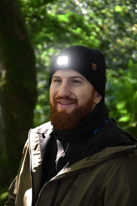 A bearded man wearing the Six Peaks LED Lighted Beanie Hat and a brown jacket smiles outdoors in a sunlit green forest.