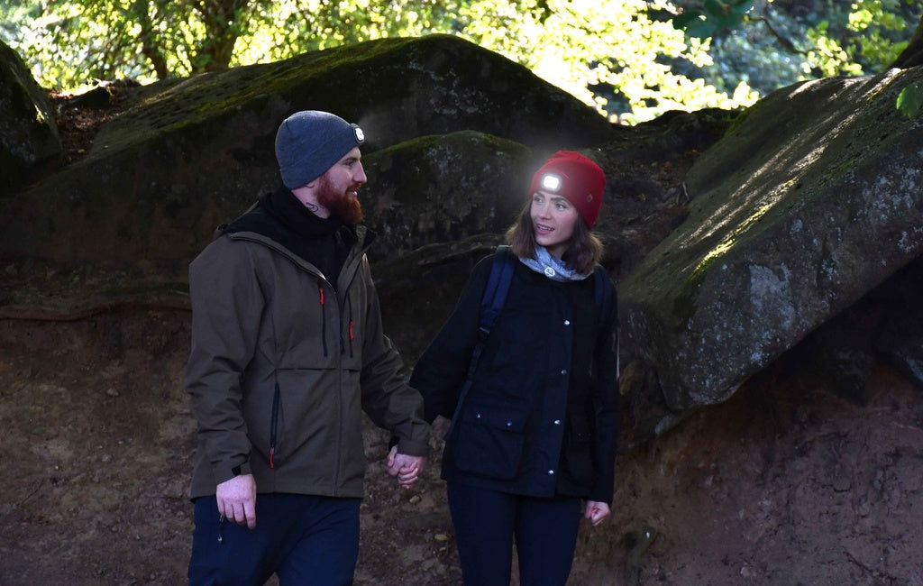 A man and woman wearing Six Peaks LED Lighted Beanie Hats hold hands while walking among large mossy rocks in the woods, bathed in natural light.