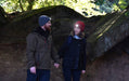 A man and woman wearing Six Peaks LED Lighted Beanie Hats hold hands while walking among large mossy rocks in the woods, bathed in natural light.