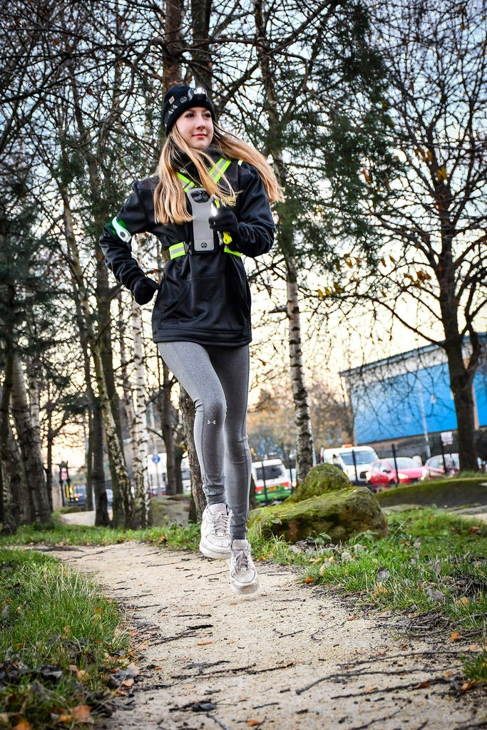 A woman in athletic wear and a beanie jogs on a park path with bare trees, wearing the Six Peaks LED Reflective Vest with Phone Holder by Six Peaks. She has a camera device on her chest, with cars and a blue building visible in the background.