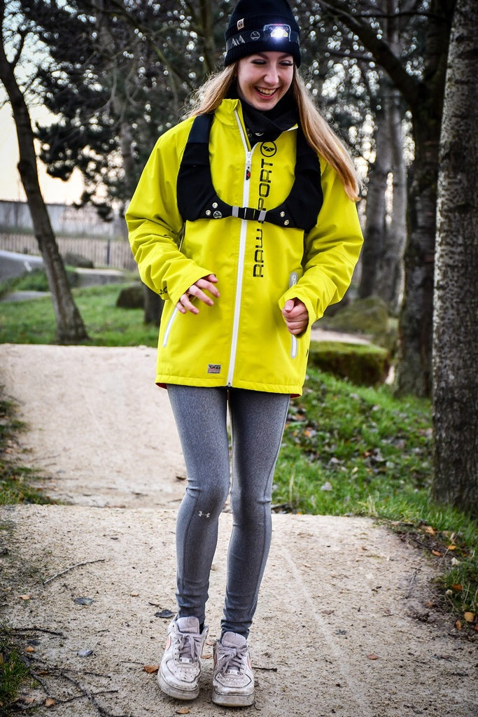 A woman smiles on a park dirt path wearing the Six Peaks LED Sport Vest, grey leggings, white sneakers, and a headlamp, with trees in the background.