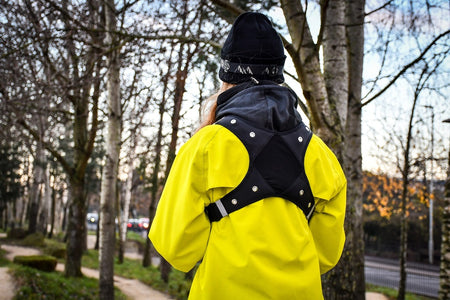 A person wearing a Six Peaks LED Sport Vest by Six Peaks and a black beanie stands on a tree-lined sidewalk in autumn or winter, seen from behind, with the harness-style vest’s reflective features visible.
