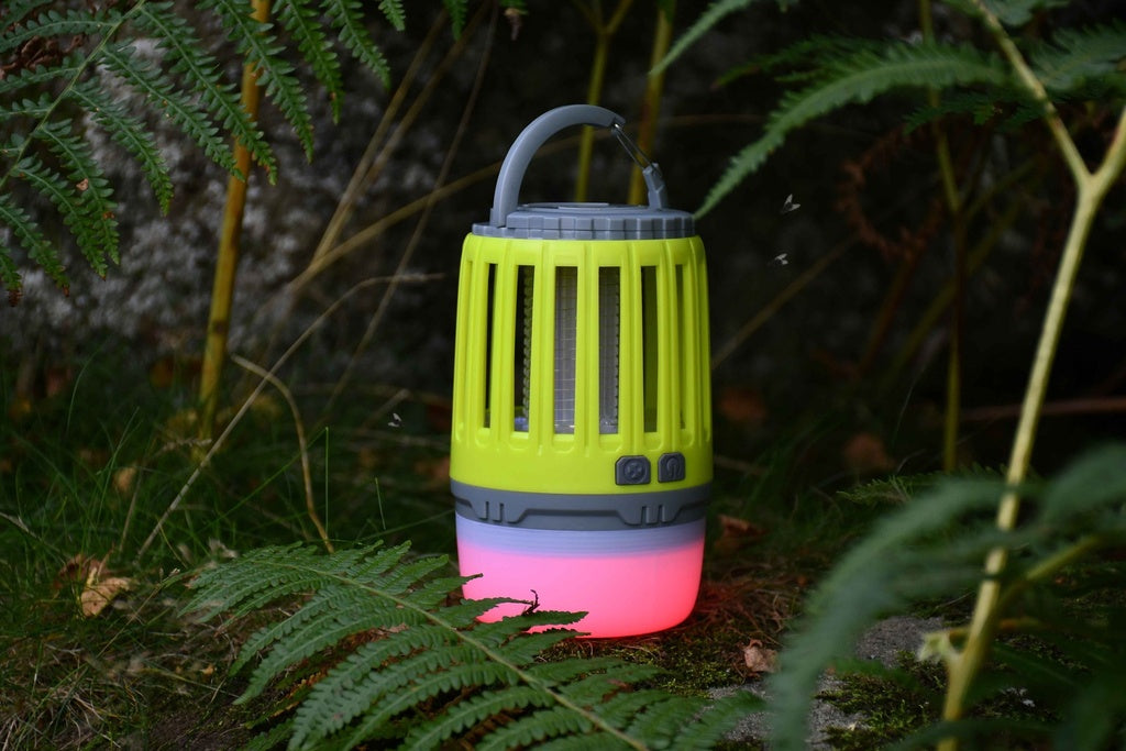 The Six Peaks Multi-function Bug Zapper Lantern by Six Peaks, featuring a green and gray design with a glowing pink base, rests on mossy ground amidst ferns outdoors—providing convenient illumination and functioning as a rechargeable bug zapper lantern for any adventure.