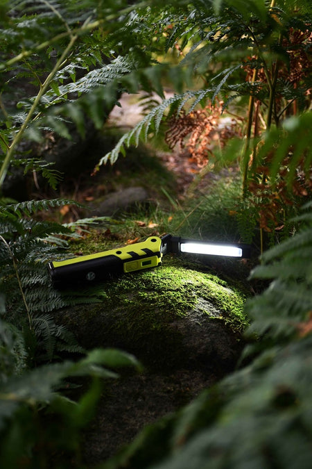 The Six Peaks Multi-function LED Work Light, featuring yellow and black accents, sits on moss among ferns in a forest, brightly illuminating its surroundings.