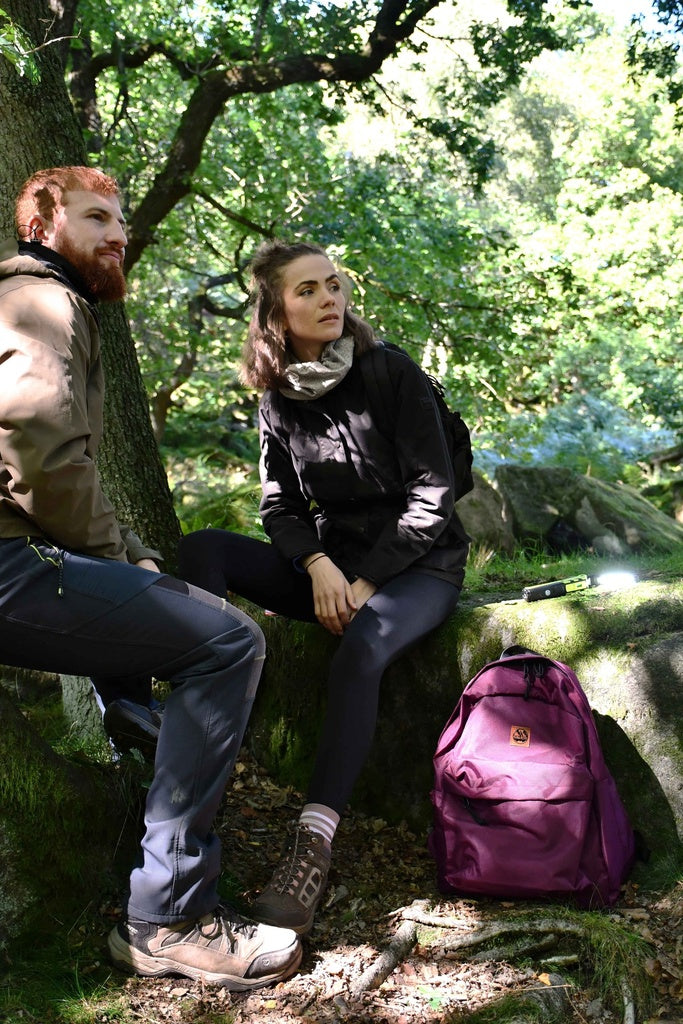 Two people in outdoor clothing sit on rocks in a sunlit forest clearing. A purple backpack and the Six Peaks Multi-function LED Work Light rest nearby, as sunlight filters through the trees, casting dappled light across the scene.