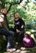 Two people in outdoor clothing sit on rocks in a sunlit forest clearing. A purple backpack and the Six Peaks Multi-function LED Work Light rest nearby, as sunlight filters through the trees, casting dappled light across the scene.
