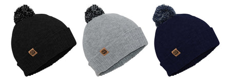 Three Six Peaks Pom Beanie winter hats are shown side by side in black, light gray, and navy blue, each with a ribbed knit design, a pom-pom on top, and a small brown logo patch on the folded brim.