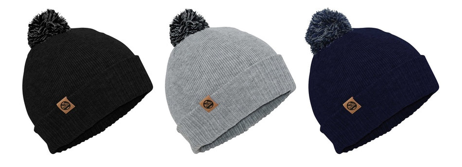Three Six Peaks Pom Beanie winter hats are shown side by side in black, light gray, and navy blue, each with a ribbed knit design, a pom-pom on top, and a small brown logo patch on the folded brim.