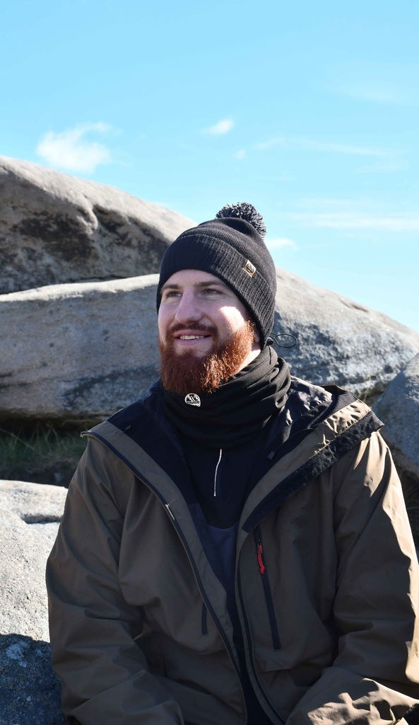A bearded man in a Six Peaks Pom Beanie by Six Peaks, paired with a dark scarf and brown jacket, smiles while sitting on large rocks under a bright blue sky.