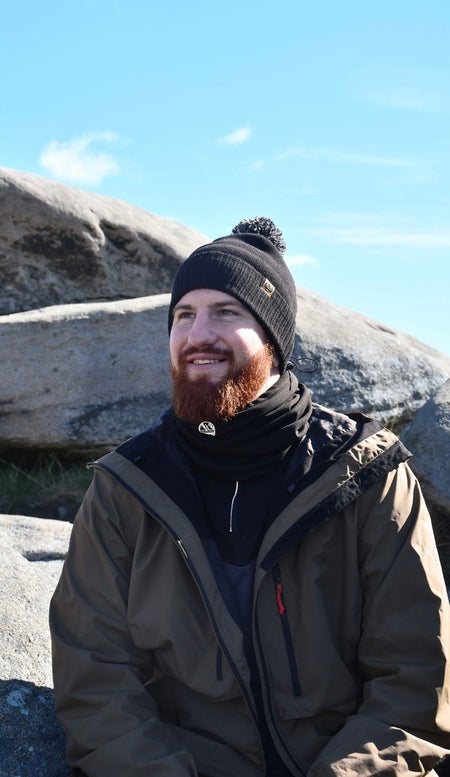 A bearded man in a Six Peaks Pom Beanie by Six Peaks, paired with a dark scarf and brown jacket, smiles while sitting on large rocks under a bright blue sky.