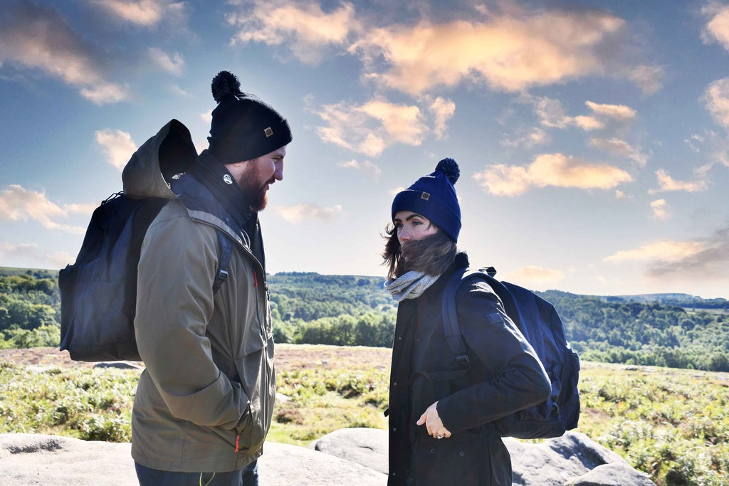 Two people wearing Six Peaks Pom Beanies and jackets stand outside with backpacks, facing each other against a scenic backdrop of greenery and rocks under a partly cloudy sunset sky.