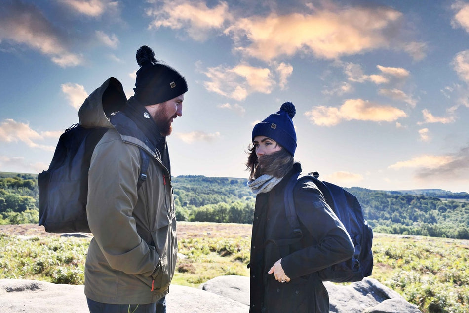 Two people wearing Six Peaks Pom Beanies and jackets stand outside with backpacks, facing each other against a scenic backdrop of greenery and rocks under a partly cloudy sunset sky.