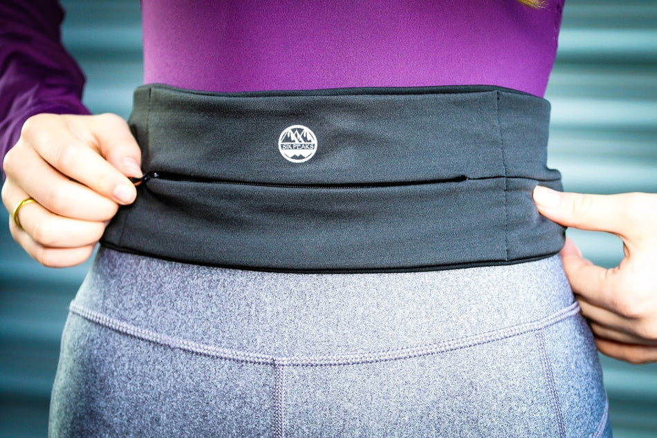 Wearing a purple top and gray leggings, a person adjusts the Six Peaks Running Slim Lightweight Waistband—a black accessory with a zipper pocket and small circular logo by Six Peaks, emphasizing its practical design.