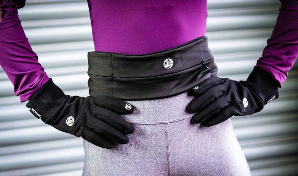 Dressed in a purple long-sleeve top, gray leggings, and black gloves, a person stands hands-on-hips before a metallic ridged wall—ready to run with their Six Peaks Running Slim Lightweight Waistband by Six Peaks.
