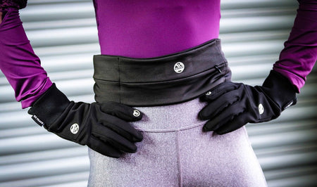 Dressed in a purple long-sleeve top, gray leggings, and black gloves, a person stands hands-on-hips before a metallic ridged wall—ready to run with their Six Peaks Running Slim Lightweight Waistband by Six Peaks.