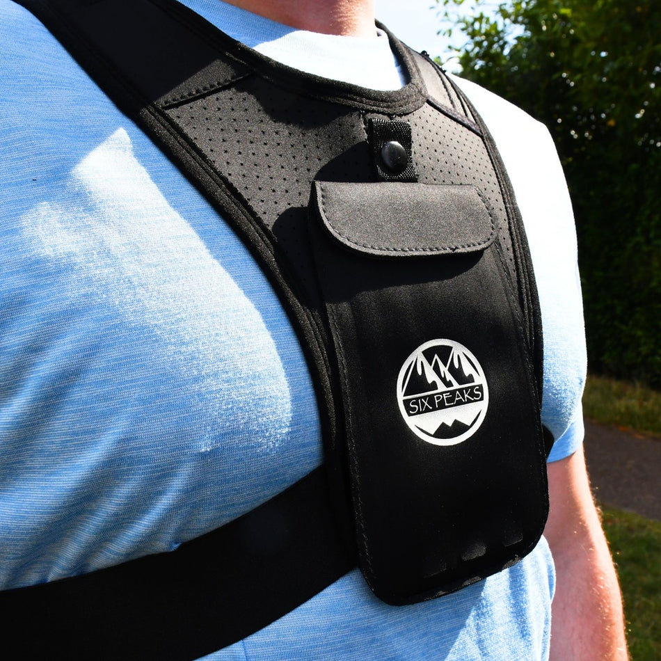 A person wears the Six Peaks Running Vest with Phone Holder, a black reflective vest from Six Peaks featuring a pocket and phone holder, over a light blue shirt outdoors with greenery in the background.