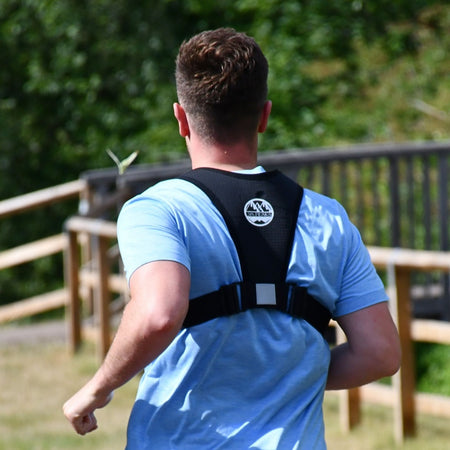 A person with short brown hair, in a light blue shirt and the Six Peaks Running Vest with Phone Holder by Six Peaks, jogs outdoors along a wooden fence surrounded by greenery.