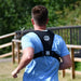 A person with short brown hair, in a light blue shirt and the Six Peaks Running Vest with Phone Holder by Six Peaks, jogs outdoors along a wooden fence surrounded by greenery.