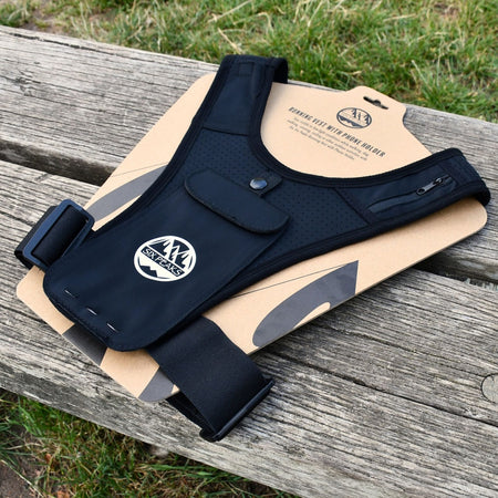 The Six Peaks Running Vest with Phone Holder by Six Peaks, in black with adjustable straps and a zippered phone pocket, is shown on cardboard packaging atop a wooden bench outdoors, with grass visible in the background.
