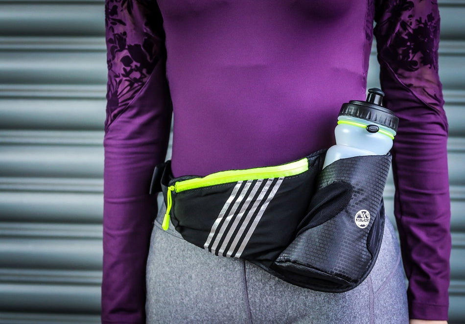A person in a purple long-sleeve top and gray pants stands by a metal shutter, wearing the Six Peaks Running Waist Belt with Bottle Holder from Six Peaks.