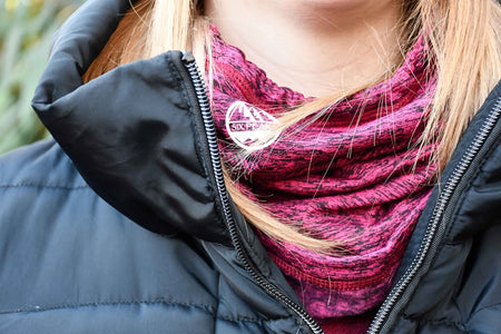 A person wearing a black puffer jacket and a maroon patterned, fleece-lined Six Peaks Winter Neck Warmer with a white Six Peaks logo, their blonde hair partially visible.