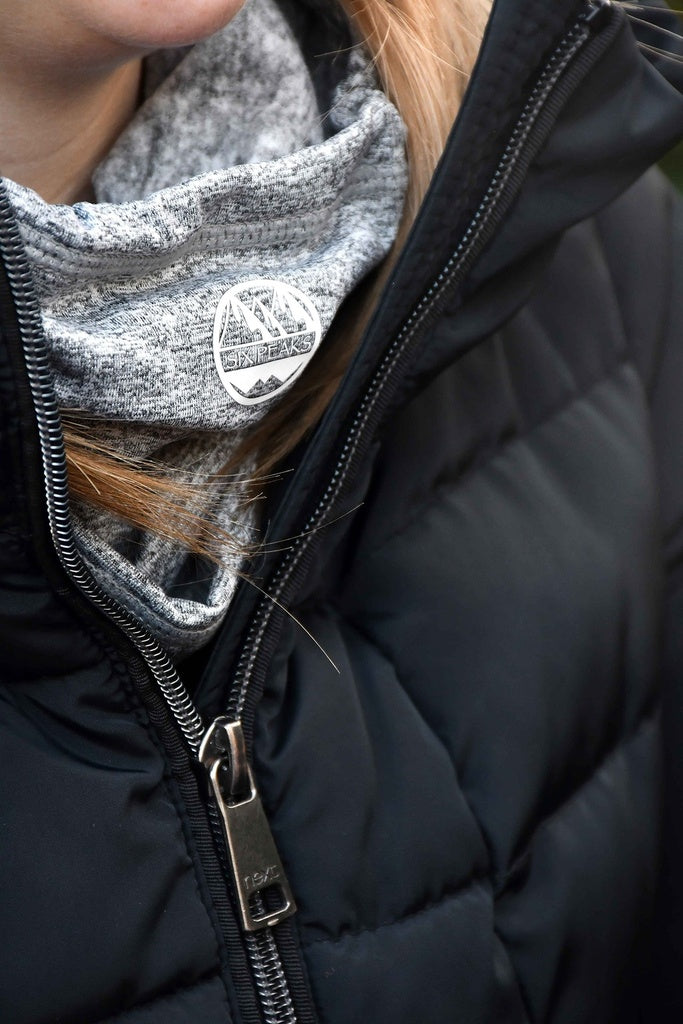 Close-up of a person wearing a black puffer jacket and the Six Peaks Winter Neck Warmer by Six Peaks, a gray wind-resistant scarf featuring a round patch with mountain graphics and the word ALASKANS. Only part of their chin and hair is visible.