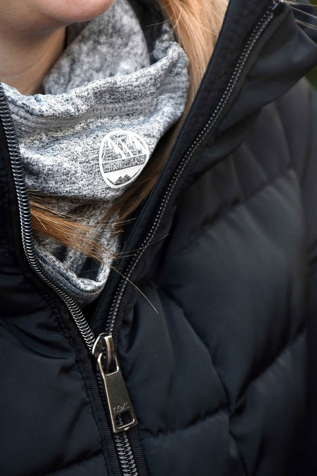 Close-up of a person wearing a black puffer jacket and the Six Peaks Winter Neck Warmer by Six Peaks, a gray wind-resistant scarf featuring a round patch with mountain graphics and the word ALASKANS. Only part of their chin and hair is visible.