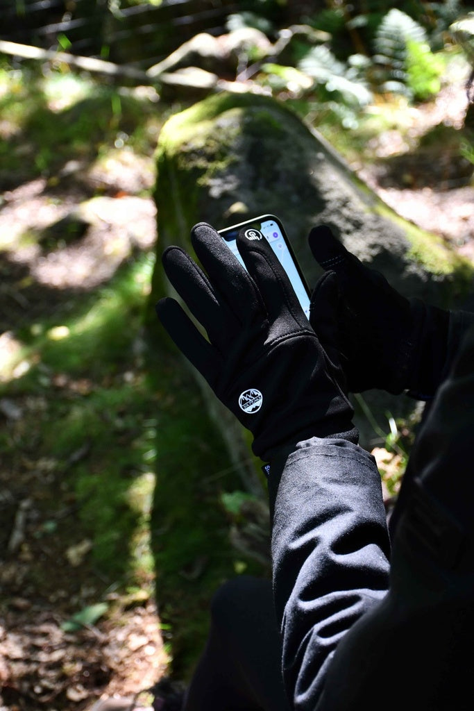 A person wearing Six Peaks Winter Thermal Gloves and a black jacket uses a smartphone outdoors, sitting on a mossy rock in a sunlit forest.