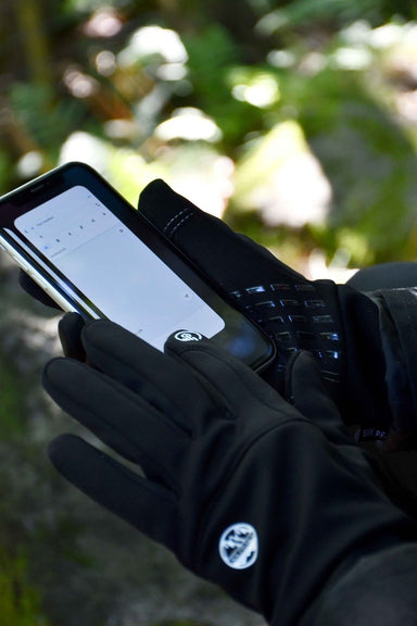 A person wearing Six Peaks Winter Thermal Gloves by Six Peaks uses a smartphone outdoors with a blank note-taking app open, while green foliage is blurred in the background.