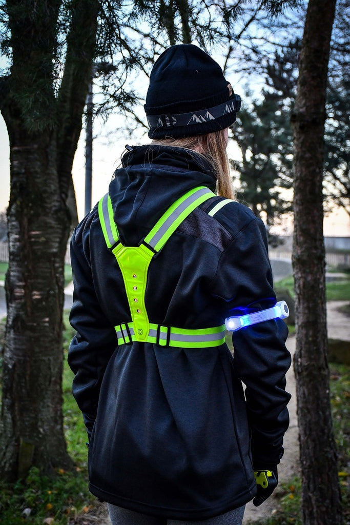A person seen from behind wears a black jacket, black hat, and bright yellow reflective harness with a Six Peaks LED Armband Running Safety Light by Six Peaks, powered by a rechargeable battery, standing outdoors among trees.