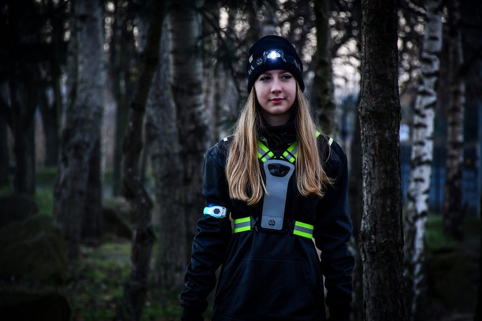 A young person stands in a misty, dimly lit forest wearing a headlamp, reflective safety gear, and a Six Peaks LED Armband Running Safety Light by Six Peaks with a rechargeable battery. Bare trees surround them as mist drifts through the eerie background.