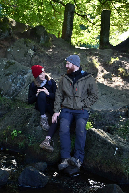 A man and woman wearing Six Peaks jackets and Six Peaks LED Lighted Beanie Hats sit on rocks by a forest stream, smiling at each other as sunlight streams through the trees, creating a peaceful, nature-filled scene.