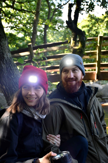 A smiling woman and man wearing Six Peaks LED Lighted Beanie Hats and outdoor jackets sit together in a wooded area, sunlight streaming through trees and a wooden bridge visible behind them.