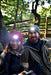 A smiling woman and man wearing Six Peaks LED Lighted Beanie Hats and outdoor jackets sit together in a wooded area, sunlight streaming through trees and a wooden bridge visible behind them.