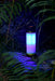 The Six Peaks Multi-function Aluminium Torch Lantern by Six Peaks, with a glowing blue light, stands on the ground amid green ferns and grass in a dark outdoor setting.