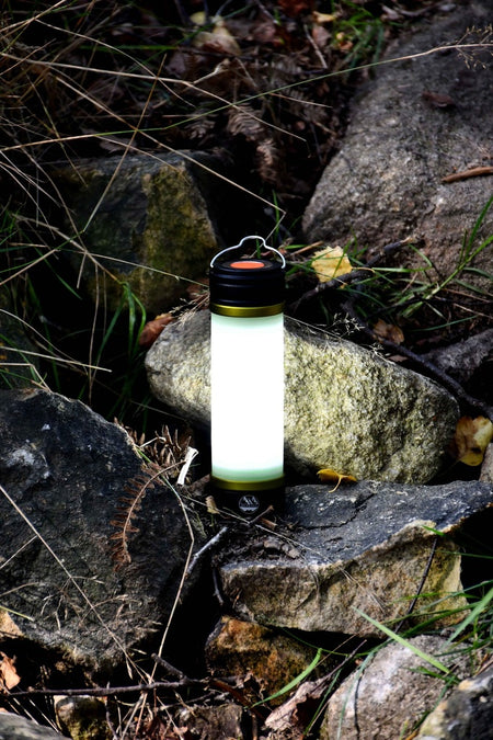 The Six Peaks Multi-function Aluminium Torch Lantern by Six Peaks sits on rocky ground amid grass and leaves, lighting up the stones around it—ideal as a camping light for any outdoor adventure.