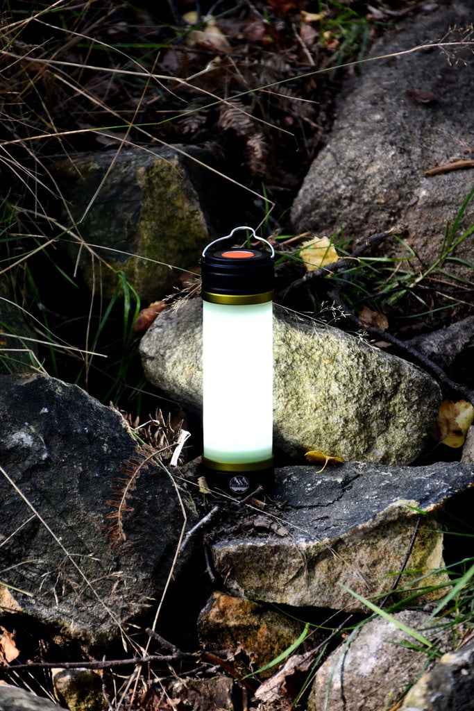 The Six Peaks Multi-function Aluminium Torch Lantern by Six Peaks sits on rocky ground amid grass and leaves, lighting up the stones around it—ideal as a camping light for any outdoor adventure.