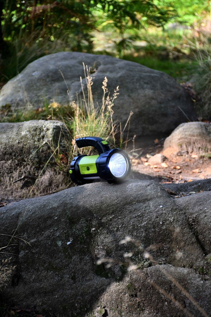 The Six Peaks Multi-function Searchlight Lantern by Six Peaks, in green and black, shines brightly on a large rock outdoors, with other rocks, grass, and foliage surrounding the portable lantern.