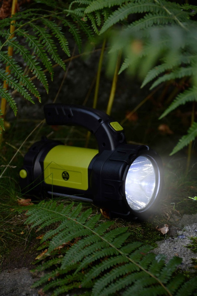 The Six Peaks Multi-function Searchlight Lantern by Six Peaks casts bright green and black LED light onto fern leaves and grass, powerfully illuminating the outdoors in a natural forest setting.