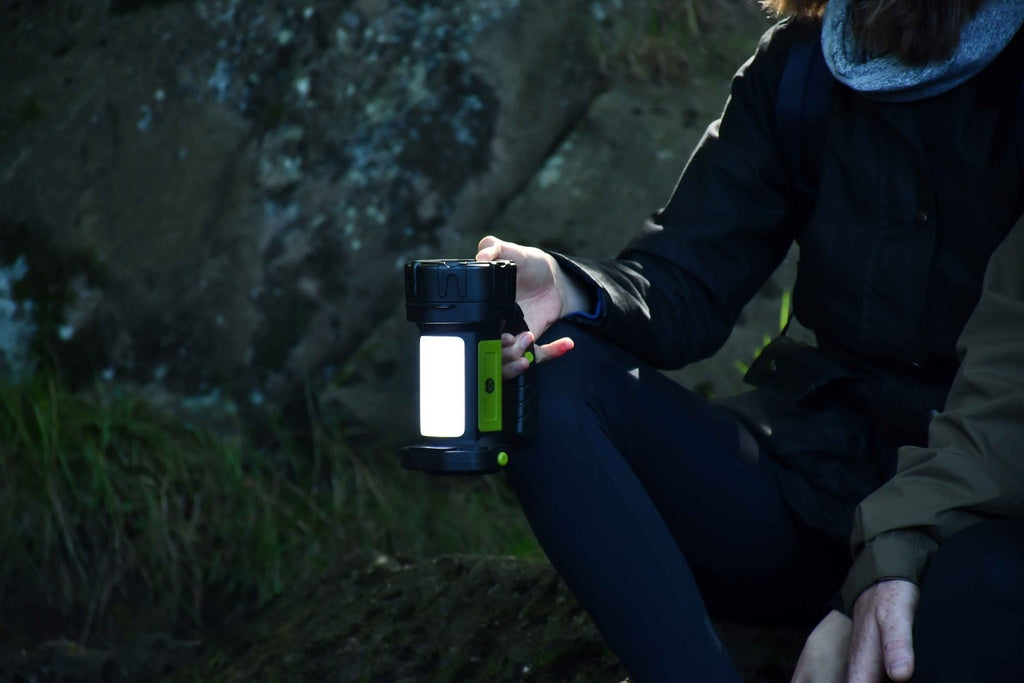 A person sitting outdoors near rocks holds a Six Peaks Multi-function Searchlight Lantern, its glow from the Six Peaks brand partially illuminating their face and surroundings in the dim natural light.