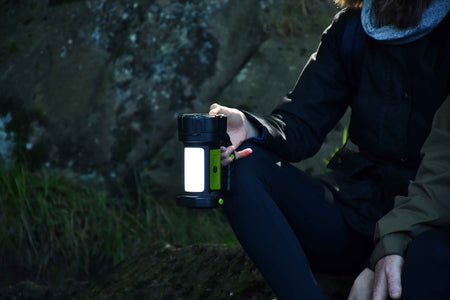 A person sitting outdoors near rocks holds a Six Peaks Multi-function Searchlight Lantern, its glow from the Six Peaks brand partially illuminating their face and surroundings in the dim natural light.