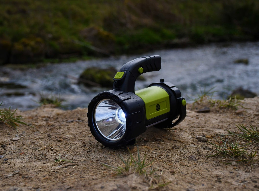 A Six Peaks Multi-function Searchlight Lantern by Six Peaks, in green and black, is switched on and resting on sandy ground near a river, with grass and water visible in the background.